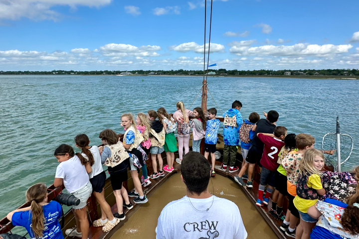Group of children on a boat with adult wearing 'crew' shirt, overlooking water under a blue sky.