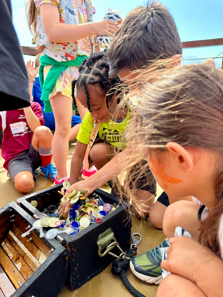 Children gather around an open treasure chest filled with coins on a wooden surface.