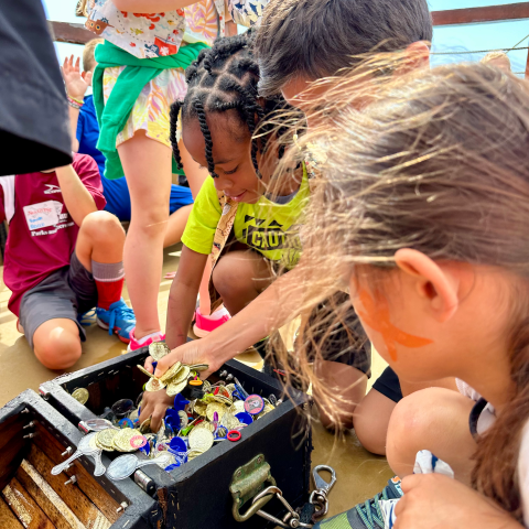 Children gather around an open treasure chest filled with coins on a wooden surface.