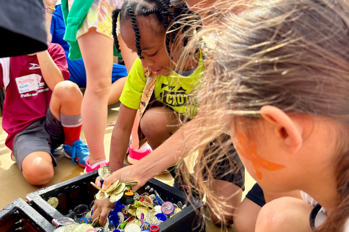 Children gather around an open treasure chest filled with coins on a wooden surface.