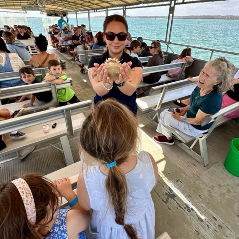 Woman on boat showing object to children, with passengers seated around.