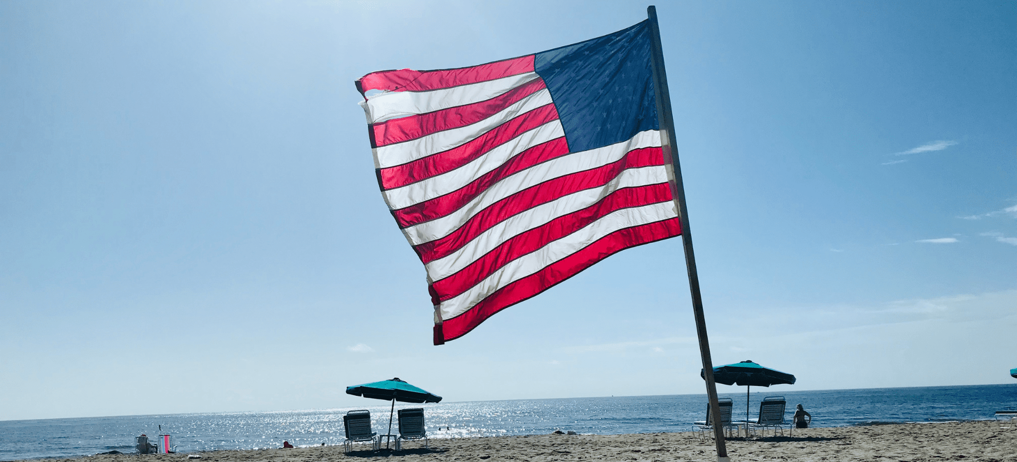 American flag waving on a beach with chairs and umbrellas, ocean in the background.