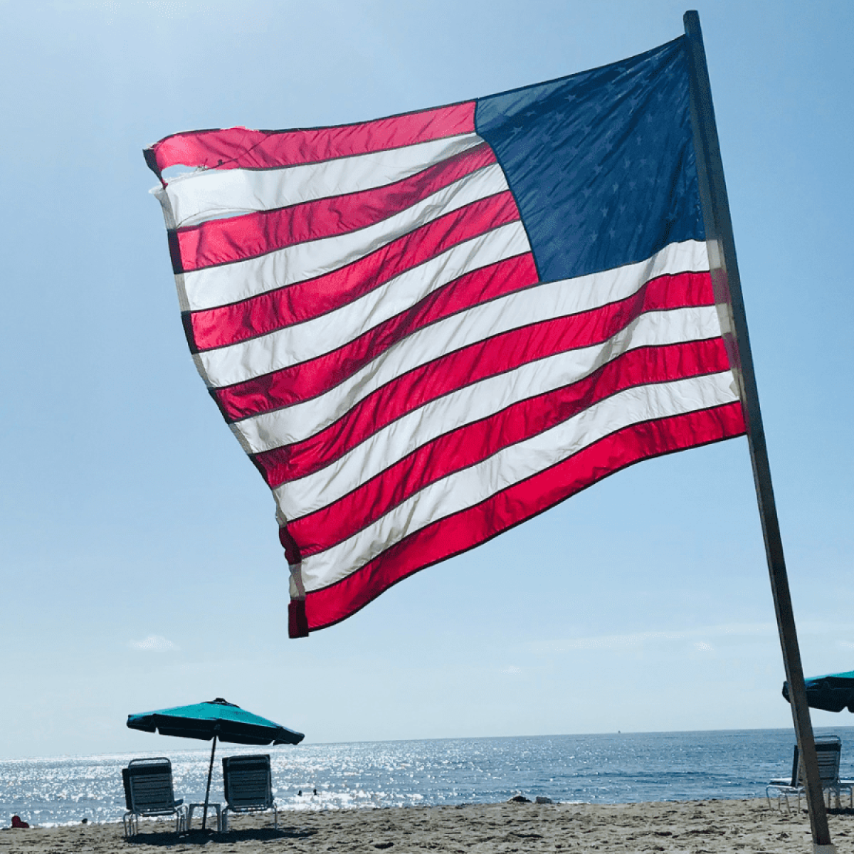 American flag waving on a beach with chairs and umbrellas, ocean in the background.
