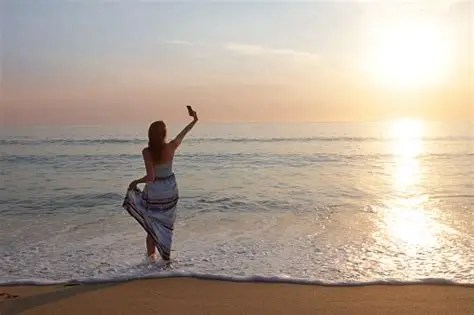 Person on beach taking a selfie with ocean and sunset in background.