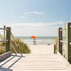 Wooden path leading to a sandy beach with a person under an orange umbrella.