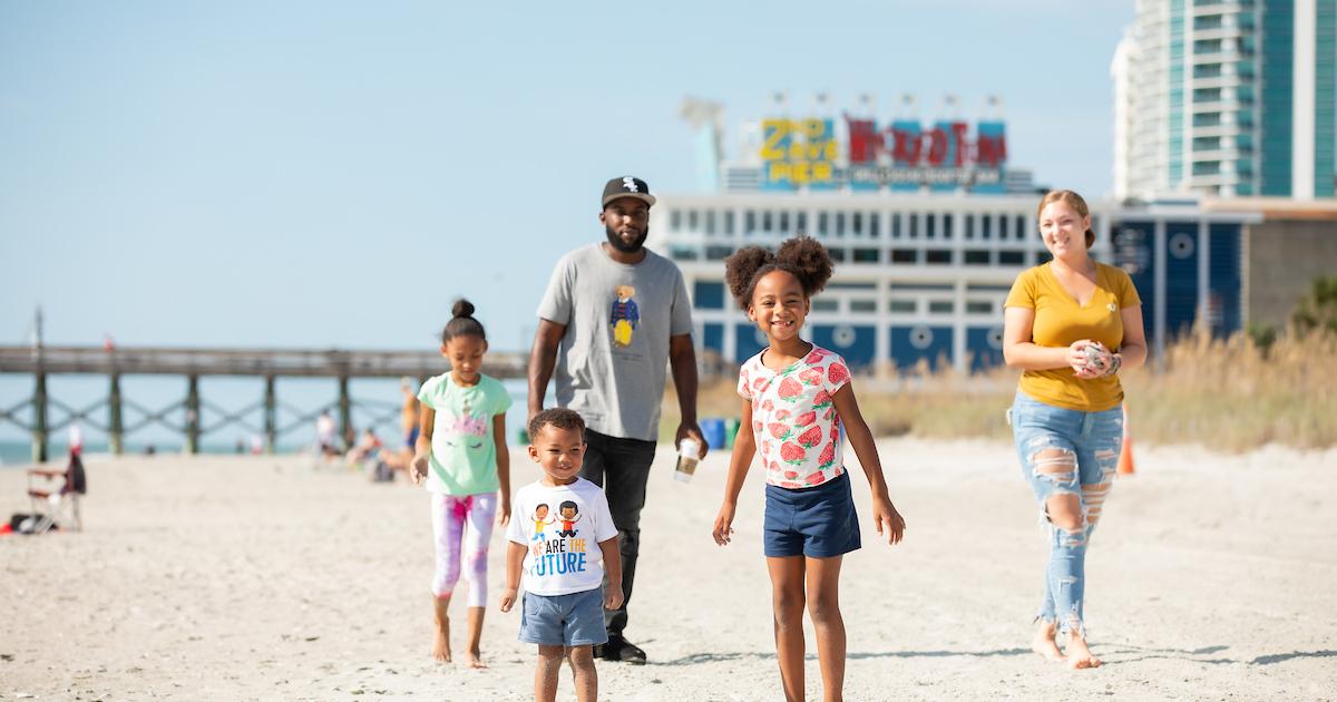 Family walking on a sunny beach with buildings in the background.