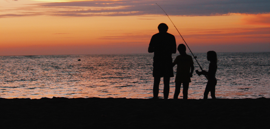 Silhouette of three people fishing on the beach at sunset.