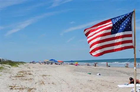 American flag on pole at sandy beach, blue sky, people in background.