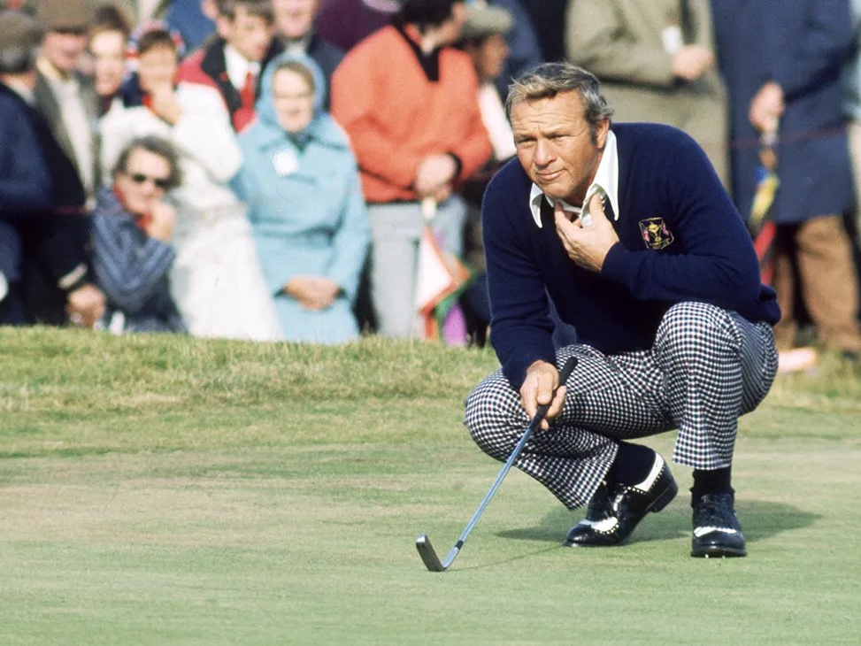 Golfer in checkered pants squatting on green, checking a putt, with spectators in background.