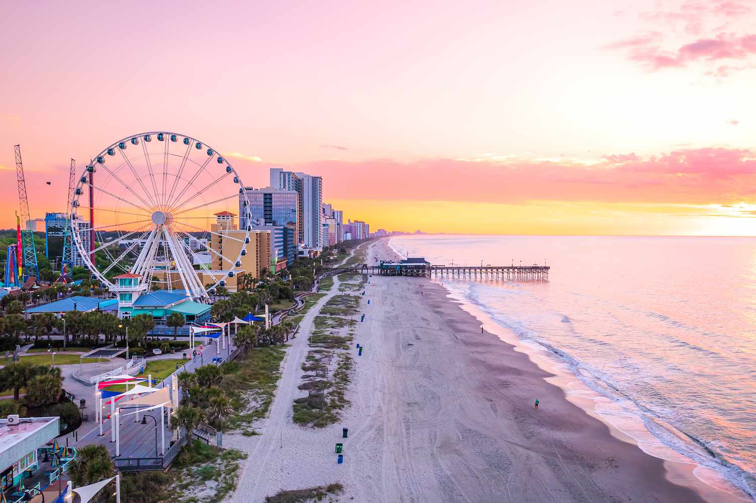 Beachfront with Ferris wheel, pier, and buildings at sunrise.