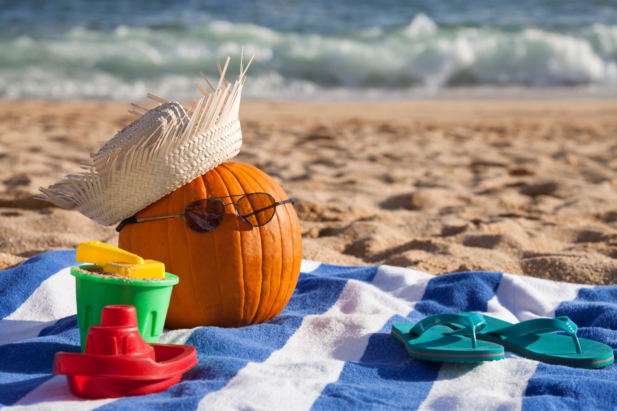 Pumpkin with hat and sunglasses on beach towel by sea.