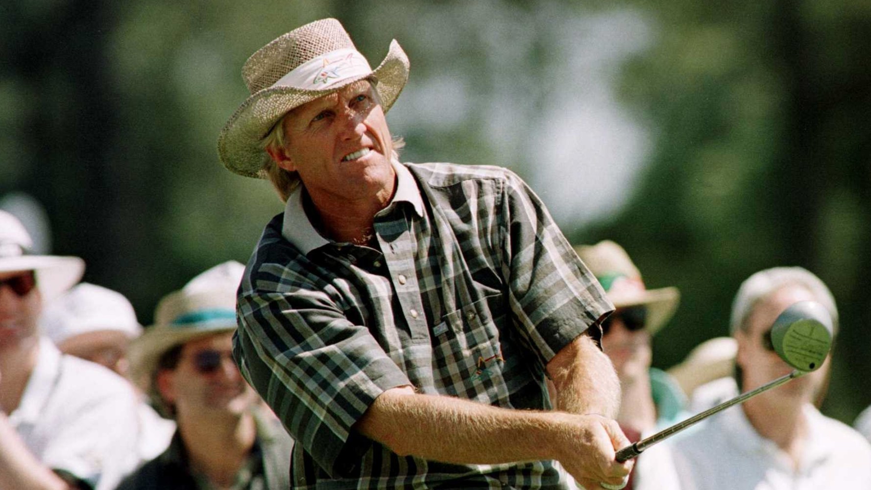 Golfer in plaid shirt and straw hat swinging a club, spectators in the background.