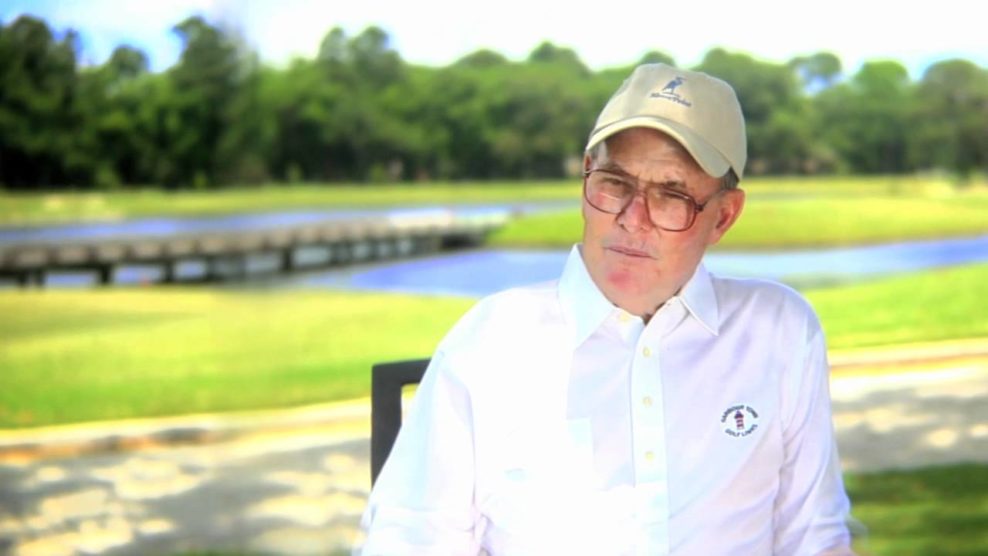 Man in a white shirt and cap sitting outdoors by a pond.