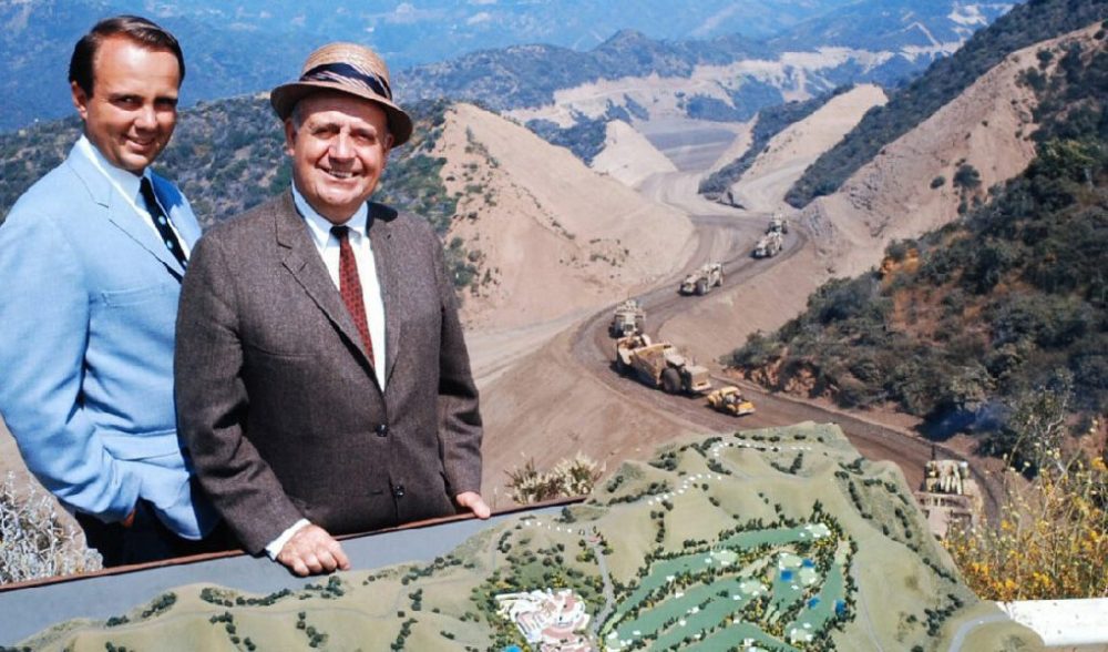 Two men in suits stand by a model overlooking a construction site in a mountainous area.