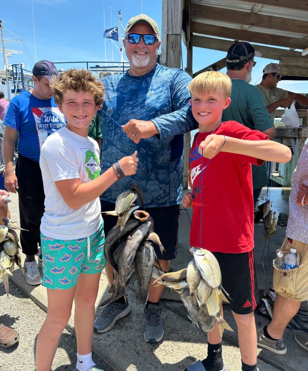 Three people holding fish on a dock, smiling, with others in the background.
