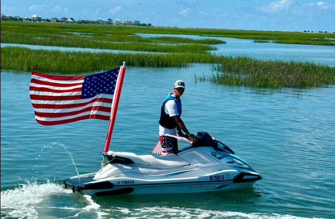 Person on jet ski with American flag on a waterway with grassy banks.