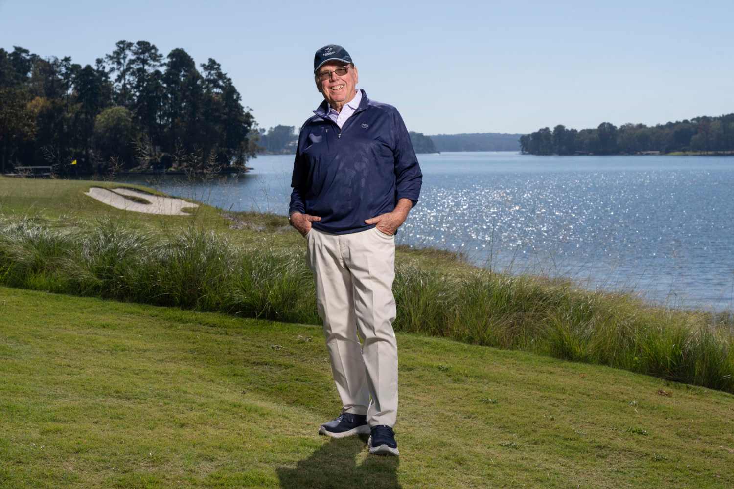 Smiling man in golf attire standing on a grassy area by a lake.