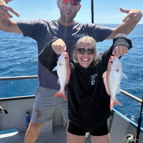 Two people on a boat holding caught fish, ocean in background.