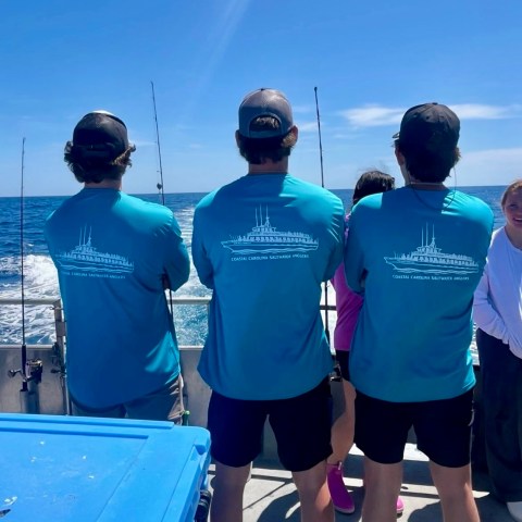 Three people on a boat fishing, wearing matching blue shirts, under a clear blue sky.