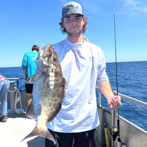 Man on boat holding a large fish with a fishing rod on a sunny day.