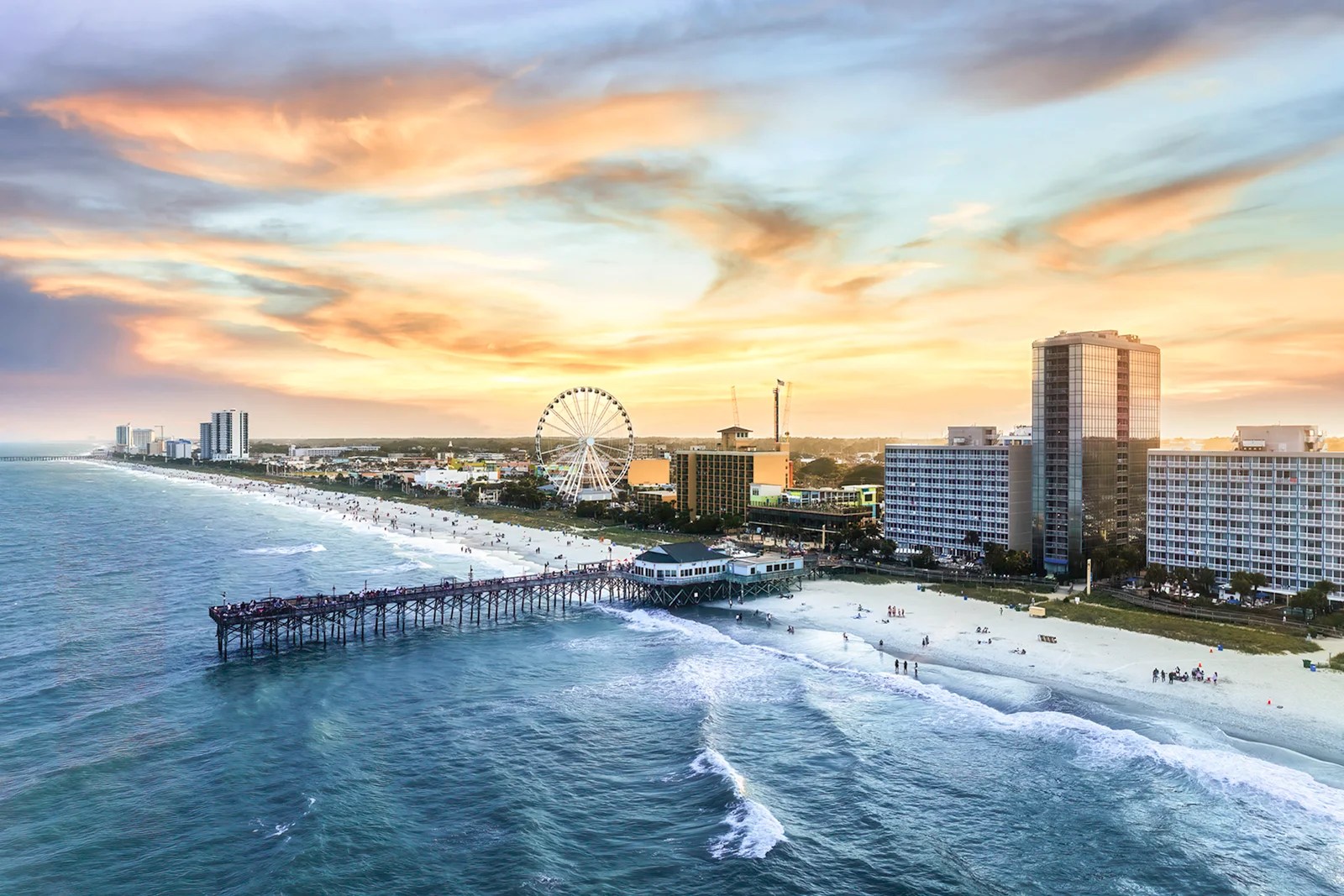 Coastal cityscape with Ferris wheel, tall buildings, pier, and sunset sky.