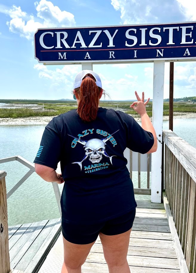 Person in black t-shirt poses with peace sign at Crazy Sister Marina sign.