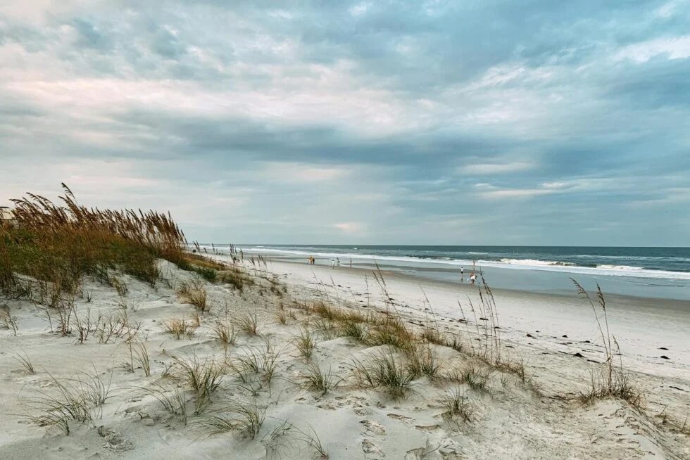 Sandy beach with sea oats and waves under a cloudy sky.