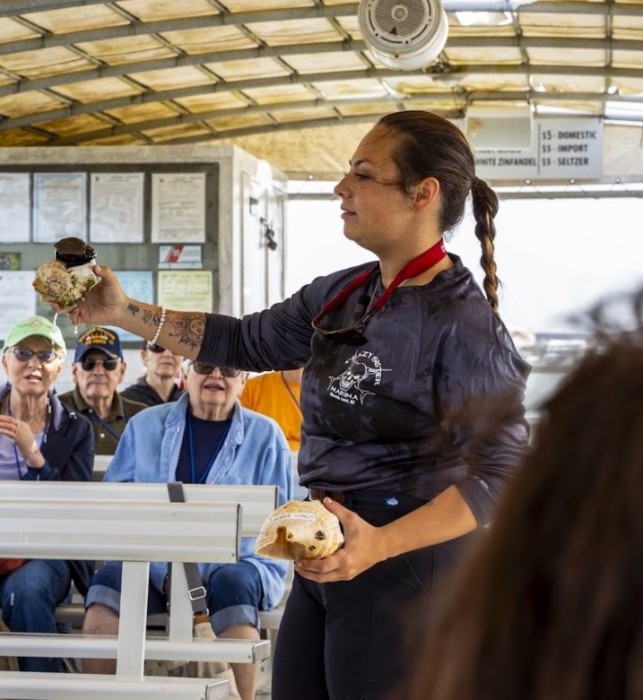 Woman holding seashells speaks to seated audience on a boat.
